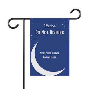 May include: A navy blue garden flag with the text "Please Do Not Disturb" and "Night Shift Worker Resting Inside". The flag features a white crescent moon and stars design. The flag is mounted on a black metal stand.