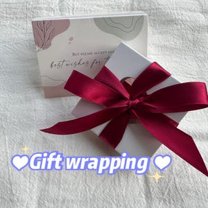 May include: A white gift box with a red satin ribbon tied in a bow. The box has a small circle cut out of the top. The box is sitting on a white surface. The text "But please accept our best wishes for..." is visible on the box.