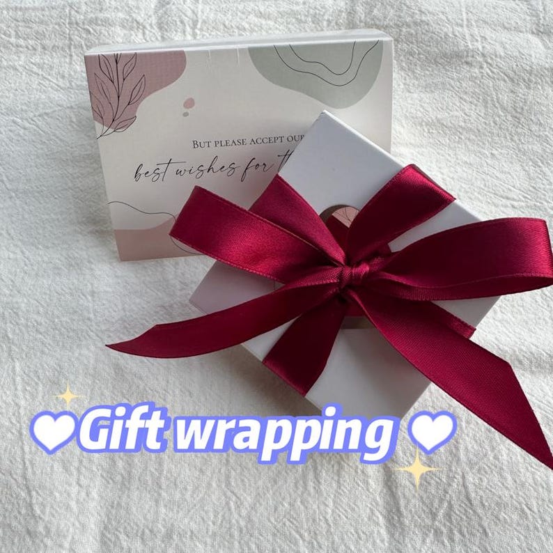 May include: A white gift box with a red satin ribbon tied in a bow. The box has a small circle cut out of the top. The box is sitting on a white surface. The text "But please accept our best wishes for..." is visible on the box.