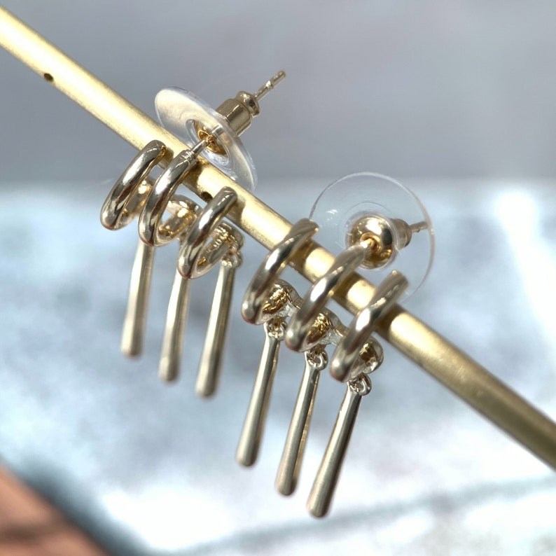 May include: Gold tone dangle earrings with multiple loops and a drop design. The earrings are displayed on a gold stand.