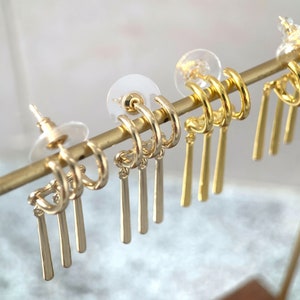 May include: Gold hoop earrings with dangling charms. The charms are thin, straight, and gold.