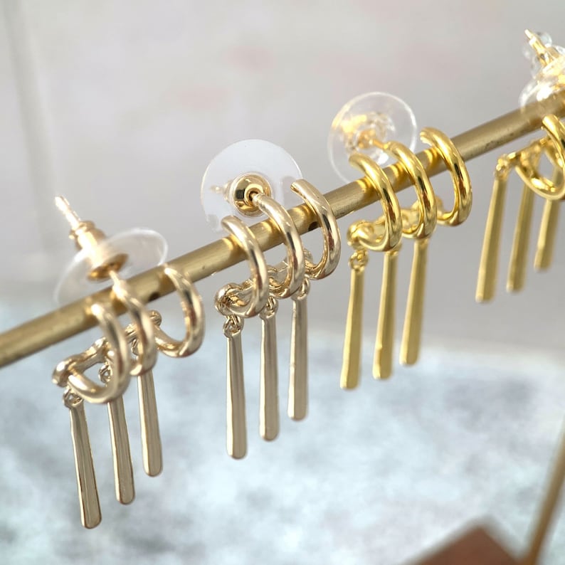 May include: Gold hoop earrings with dangling charms. The charms are thin, straight, and gold.