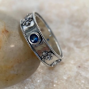 Könnte beinhalten: Ein silberner Bandring mit einem blauen Edelstein und floralen Designs.