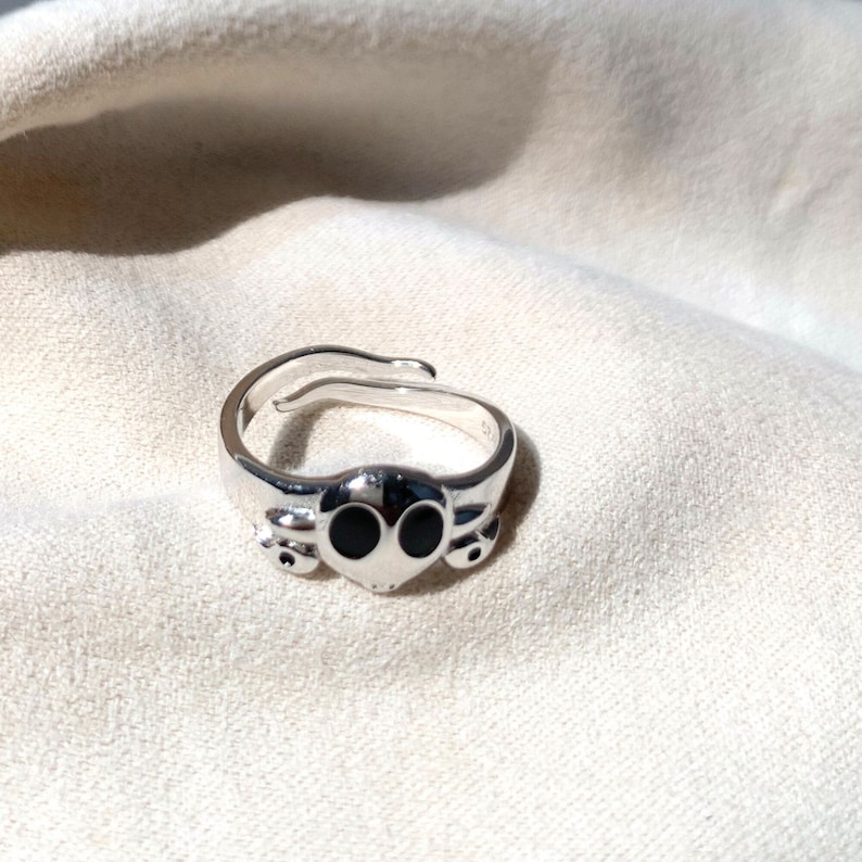 K&ouml;nnte beinhalten: Ein silberner Ring mit einem schwarzen Alienkopf-Design. Der Ring ist verstellbar und hat einen schlichten, modernen Stil.