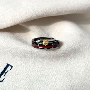 K&ouml;nnte beinhalten: Ein schwarzer Ring mit einer roten Linie und kleinen goldenen Akzenten. Der Ring hat ein strukturiertes Design und ist mit kleinen klaren Steinen besetzt.