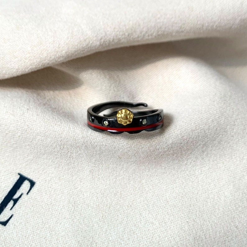 K&ouml;nnte beinhalten: Ein schwarzer Ring mit einer roten Linie und kleinen goldenen Akzenten. Der Ring hat ein strukturiertes Design und ist mit kleinen klaren Steinen besetzt.