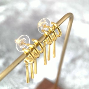 May include: Gold hoop earrings with three dangling charms. The hoops are stacked on top of each other and the charms are thin and cylindrical.