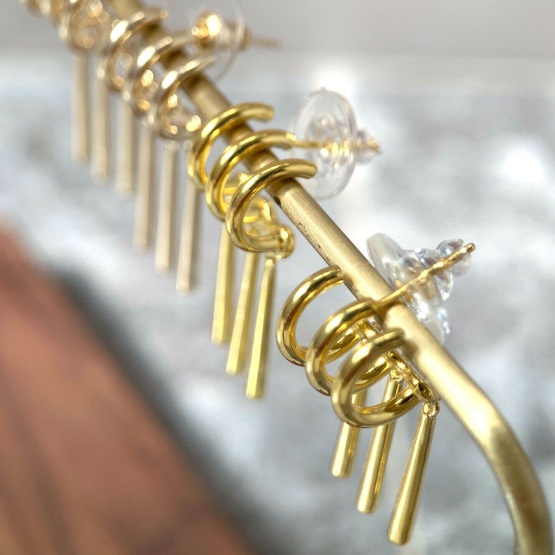 May include: Gold hoop earrings with dangling gold bars. The earrings are displayed on a gold stand.