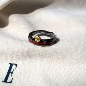 K&ouml;nnte beinhalten: Ein schwarzer Ring mit einem roten Band und einem goldenen Akzent. Der Ring ist mit kleinen schwarzen Steinen besetzt.