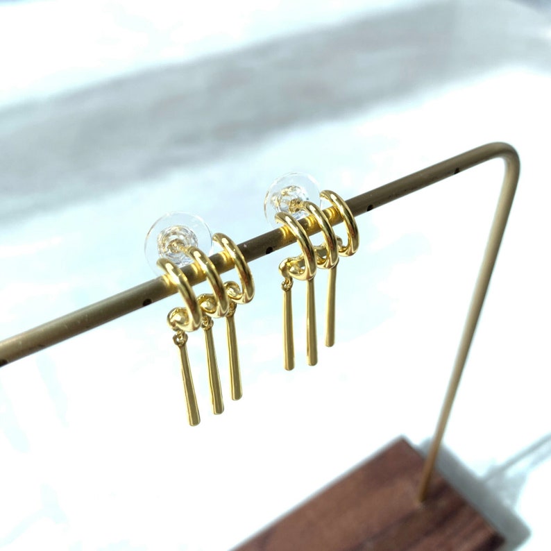 May include: Gold hoop earrings with three dangling chains. The earrings are on a display stand.