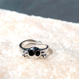 K&ouml;nnte beinhalten: Ein silberner Ring mit einem Totenkopfdesign mit zwei schwarzen Augen. Der Ring ist verstellbar und hat einen schlichten, minimalistischen Stil.