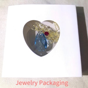May include: A white heart-shaped jewelry box with a blue teardrop-shaped pendant necklace inside. The necklace has a gold chain and a red gemstone accent.