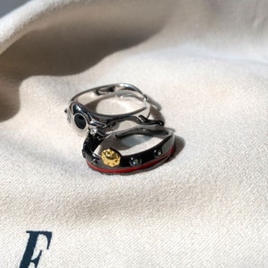 K&ouml;nnte beinhalten: Zwei silberne Ringe mit schwarzen und roten Akzenten. Der obere Ring hat einen schwarzen Stein mit einer silbernen Fassung. Der untere Ring hat ein rotes Band mit schwarzen Steinen und einem goldenen Akzent.