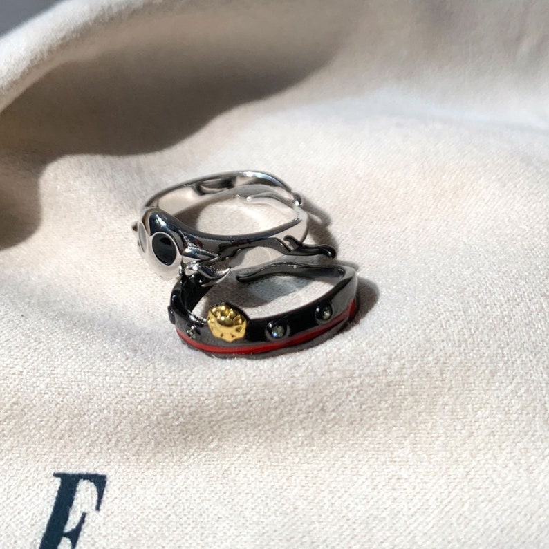K&ouml;nnte beinhalten: Zwei silberne Ringe mit schwarzen und roten Akzenten. Der obere Ring hat einen schwarzen Stein mit einer silbernen Fassung. Der untere Ring hat ein rotes Band mit schwarzen Steinen und einem goldenen Akzent.