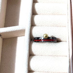K&ouml;nnte beinhalten: Ein schwarzer Ring mit einem roten Band und kleinen klaren Steinen. Der Ring hat oben ein goldenes Blumendesign.