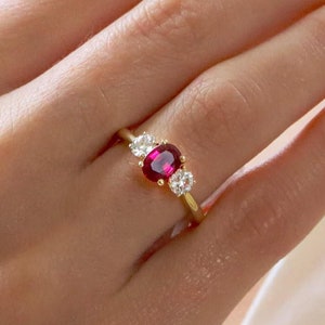 May include: A gold ring with a ruby and two diamonds. The ruby is oval shaped and the diamonds are round. The ring is on a person's finger.