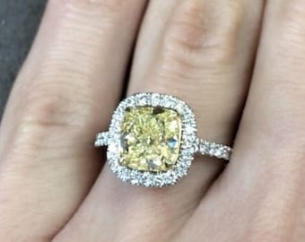 May include: A diamond ring with a large, square, yellow center stone, surrounded by a halo of smaller diamonds. The ring is set in a silver band, and the hand has burgundy nail polish.