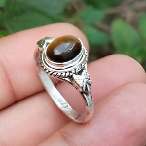 Könnte beinhalten: Ein silberner Ring mit einem ovalen, braunen Tigeraugenstein. Der Ring hat ein verziertes Design um den Stein und am Band. Der Text "Silver Jewellery Exports" befindet sich am unteren Rand des Bildes.
