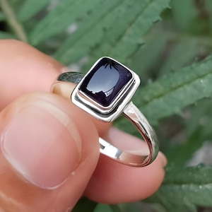Puede incluir: Un anillo de plata con una piedra preciosa cuadrada de color púrpura oscuro. El anillo se muestra sobre un fondo de hojas verdes. El texto "Silver Jewellery Exports" es visible en la parte inferior de la imagen.