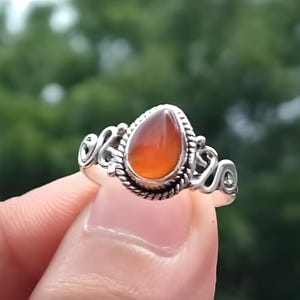 May include: A silver ring featuring a teardrop-shaped, orange gemstone. The ring has an ornate, swirling design around the stone and the band. The text "Silver Jewellery Exports" is visible at the top.