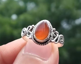 May include: A silver ring featuring a teardrop-shaped, orange gemstone. The ring has an ornate, swirling design around the stone and the band. The text "Silver Jewellery Exports" is visible at the top.