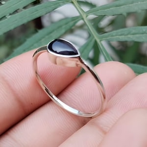 May include: A silver ring with a teardrop-shaped black gemstone. The ring is held in a hand against a backdrop of green foliage. The text "Silver Jewellery Exports" is visible at the bottom of the image.