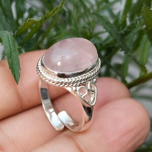 May include: A silver ring with a large, oval rose quartz gemstone. The ring has a braided border around the stone and decorative openwork on the band. The text "Silver Jewellery Exports" is at the bottom of the image.