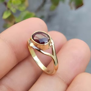 May include: A gold ring featuring a dark red oval gemstone. The ring has a distinctive design with a split band that curves around the stone. The text "Silver Jewellery Exports" is visible in the background.