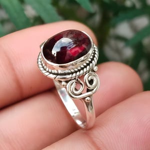 Peut inclure: Une bague en argent ornée d'une pierre précieuse ovale rouge foncé. La pierre est sertie dans une lunette détaillée avec un motif de corde torsadée. La bande de la bague présente un motif tourbillonnant orné. Le texte "Silver Jewellery Exports" est en bas.