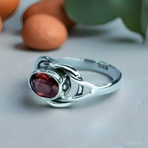 Può includere: Un anello d'argento con una gemma ovale rosso scuro. L'anello presenta un design unico con elementi metallici intrecciati. Il testo "SILVER JEWELLERY EXPORTS" è in basso nell'immagine.