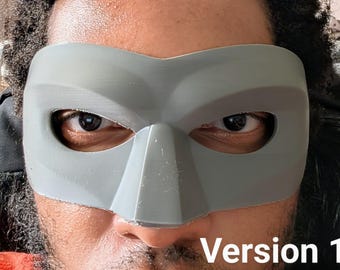 Peut inclure: Un masque gris imprimé en 3D avec un nez pointu et des ouvertures pour les yeux. Le masque couvre le haut du visage, avec le texte "Version 1" en bas. Le masque est porté par une personne.