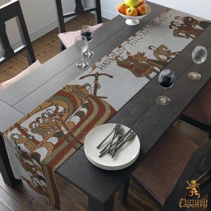May include: A long rectangular table runner featuring a woven design inspired by the Bayeux Tapestry, displaying historical figures in earth tones of brown, green, and red on a light background with the text HAROLD: HIC: APPREHENDIT.
