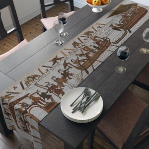 May include: A beige and brown tapestry table runner with a historical illustration depicting a boat and figures. The runner is placed on a dark wooden table, alongside white plates, silverware, and wine glasses.