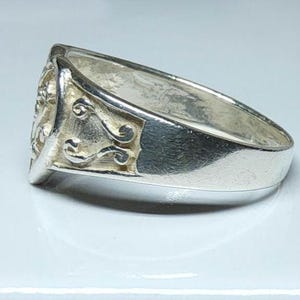 Puede incluir: Anillo de plata con un dise&ntilde;o detallado en la cara y adornos decorativos en los laterales. El anillo tiene una banda ancha y un acabado pulido, que refleja la luz.
