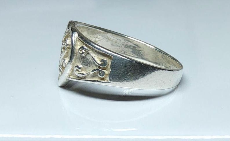 Puede incluir: Anillo de plata con un dise&ntilde;o detallado en la cara y adornos decorativos en los laterales. El anillo tiene una banda ancha y un acabado pulido, que refleja la luz.