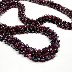 May include: A long strand of deep red garnet beads, featuring numerous small, faceted, teardrop-shaped stones strung together. The necklace displays a rich, dark burgundy color with a reflective, polished surface across the many individual beads.