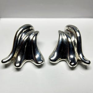 May include: A pair of polished silver-tone earrings featuring a fluid, sculptural design with smooth, undulating curves that create a sense of movement. The metallic surface is highly reflective, highlighting the organic, wave-like shape of each piece.