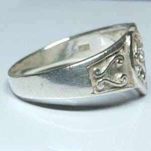 Puede incluir: Un anillo de plata con una cara rectangular con un dise&ntilde;o ornamental en espiral. El anillo tiene una banda lisa y pulida y una superficie ligeramente texturizada. El anillo se muestra sobre una superficie blanca.