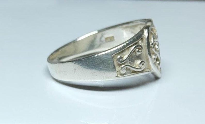 Puede incluir: Un anillo de plata con una cara rectangular con un dise&ntilde;o ornamental en espiral. El anillo tiene una banda lisa y pulida y una superficie ligeramente texturizada. El anillo se muestra sobre una superficie blanca.