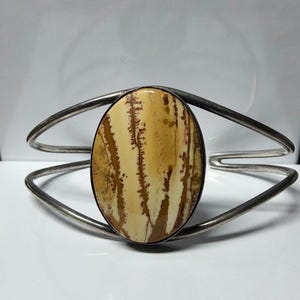 Può includere: Bracciale a polsino argentato con una pietra ovale color crema con venature marroni e beige. La pietra è incastonata in una lunetta nera. Il bracciale ha un design minimalista con una fascia divisa che si curva attorno al polso.