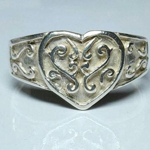 Puede incluir: Un anillo de plata con un coraz&oacute;n en el centro. El coraz&oacute;n est&aacute; lleno de un dise&ntilde;o ornamental y arremolinado. La banda del anillo presenta un patr&oacute;n de desplazamiento repetido. El anillo est&aacute; sobre un fondo blanco.