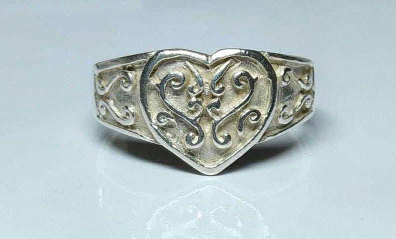 Puede incluir: Un anillo de plata con un coraz&oacute;n en el centro. El coraz&oacute;n est&aacute; lleno de un dise&ntilde;o ornamental y arremolinado. La banda del anillo presenta un patr&oacute;n de desplazamiento repetido. El anillo est&aacute; sobre un fondo blanco.