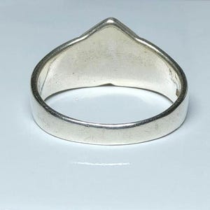Puede incluir: Un anillo de plata con una parte superior triangular. El anillo tiene una banda lisa y pulida y una superficie plana y sin adornos. El anillo est&aacute; sobre un fondo blanco.