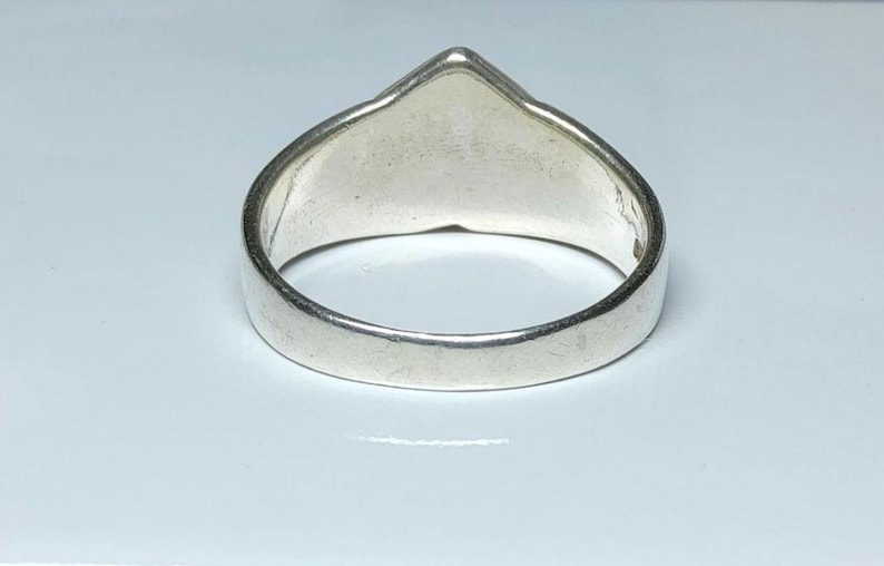 Puede incluir: Un anillo de plata con una parte superior triangular. El anillo tiene una banda lisa y pulida y una superficie plana y sin adornos. El anillo est&aacute; sobre un fondo blanco.