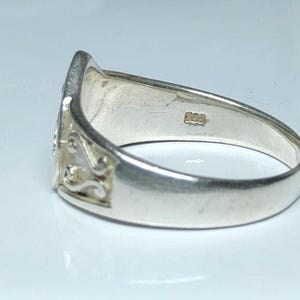 Puede incluir: Un anillo de plata con un dise&ntilde;o ornamentado en la cara. El anillo tiene una banda lisa y pulida y un elemento decorativo con un patr&oacute;n floral o de volutas. El anillo est&aacute; sobre un fondo blanco.