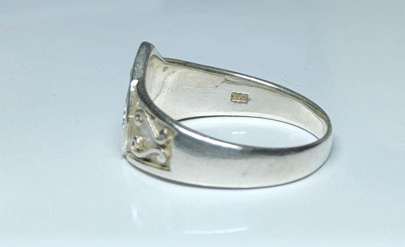 Puede incluir: Un anillo de plata con un dise&ntilde;o ornamentado en la cara. El anillo tiene una banda lisa y pulida y un elemento decorativo con un patr&oacute;n floral o de volutas. El anillo est&aacute; sobre un fondo blanco.