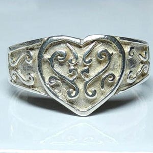 Puede incluir: Anillo de plata con dise&ntilde;o en forma de coraz&oacute;n. El coraz&oacute;n es el punto focal, con un intrincado patr&oacute;n arremolinado. La banda tiene un dise&ntilde;o decorativo de volutas. El anillo est&aacute; sobre un fondo claro.