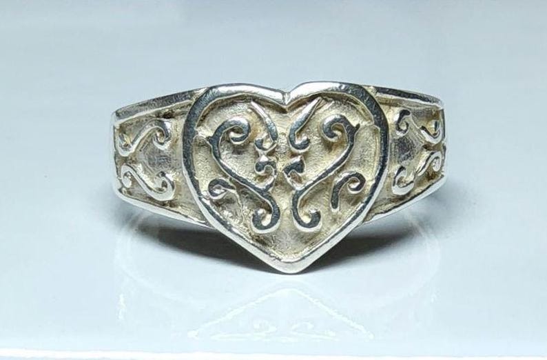 Puede incluir: Anillo de plata con dise&ntilde;o en forma de coraz&oacute;n. El coraz&oacute;n es el punto focal, con un intrincado patr&oacute;n arremolinado. La banda tiene un dise&ntilde;o decorativo de volutas. El anillo est&aacute; sobre un fondo claro.
