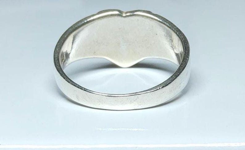 Puede incluir: Un anillo de plata con un dise&ntilde;o en forma de coraz&oacute;n. El anillo tiene una banda ancha y una superficie lisa y pulida. La forma de coraz&oacute;n es visible en la parte superior del anillo.