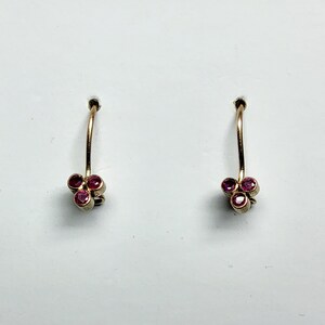 Puede incluir: Un par de pendientes colgantes de oro, cada uno con un grupo de tres pequeños rubíes redondos de color rosa intenso dispuestos en forma triangular en la base de un gancho de alambre de oro curvado.